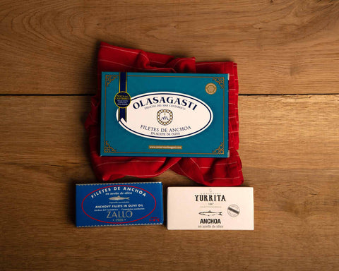 One large blue tin of Olasagasti anchovies on a red tea towel, with two smaller tins of anchovies in blue and white packaging beneath it