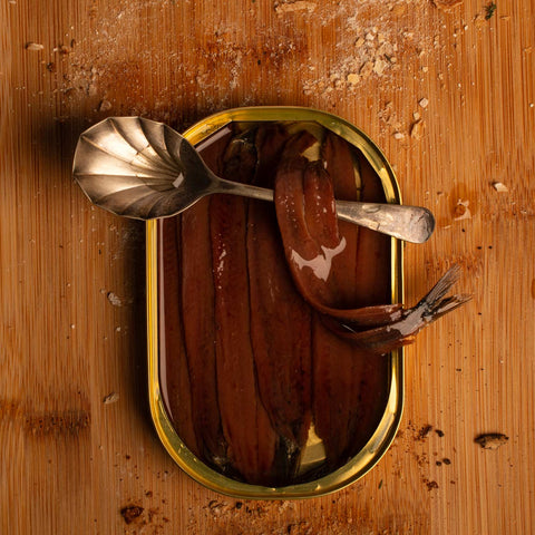 Tinned anchovies in olive oil on a wooden backdrop scattered with crumbs. With a silver spoon at the ready. 