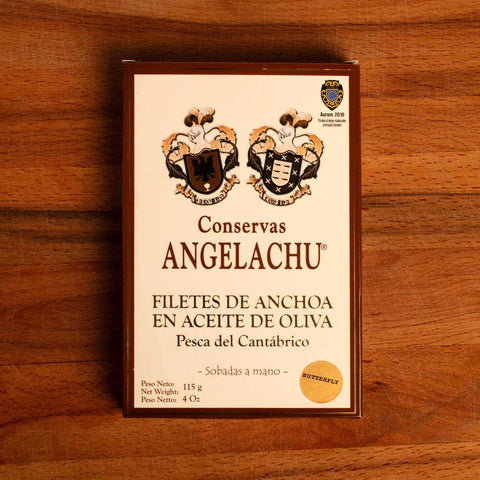 A tin of butterfly anchovies in white card packaging with brown borders, featuring the text "Conservas Angelachu" on the front.