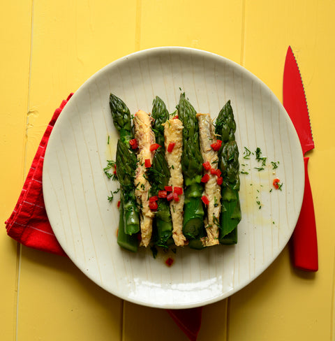 Pale mackerel fillets placed between vibrant green spears of asparagus and sprinkled with finely diced red chilli and chopped dill. On a clear plate with a red napkin and knife, all sitting comfortably on a yellow wooden table.