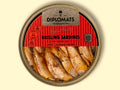 A circular golden-coloured tin of brisling sardines or sprats. Half of the lid is clear. Through it you can see the neatly arranged sprats in an orange-tinged oil. The upper half of the lid is red with Diplomats written in black lettering at the top of this against a golden background. The tin is on a pale yellow background.