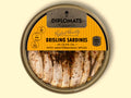 A circular golden-coloured tin of brisling sardines or sprats. Half of the lid is clear. Through it you can see the neatly arranged sprats in oil with some spices gathered towards the centre. The upper half of the lid is yellow with Diplomats written in black lettering at the top of this against a golden background. The tin is against a pale yellow background.