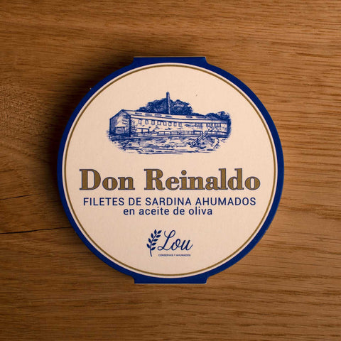 A round tin with a white card wrap. The cover has a blue and gold border. Don Reinaldo is written in gold lettering below a drawing of the cannery.