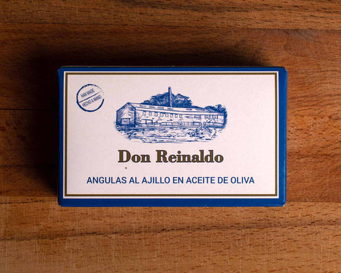 A tin in white packaging with a blue border with a picture of Don Reinaldo cannery on the front. The tin is on a wood background.