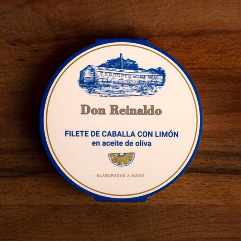 A round tin of mackerel fillets with lemon, with a wrap made of white card and a blue and white and gold border. Don Reinaldo is written in gold lettering on the front below a drawing of the cannery.