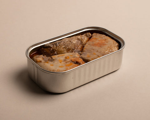 An open silver rectangular tin of monkfish liver preserved in its own oil, set against a grey backdrop.