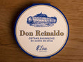 A round tin with a white card wrap. The cover has a blue and gold border. Don Reinaldo is written in gold lettering below a drawing of the cannery.