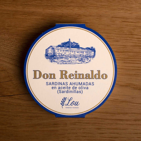 A round tin with a white card wrap. The cover has a blue and gold border. Don Reinaldo is written in gold lettering below a drawing of the cannery.