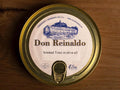 A round gold-coloured tin with a sticker on the front. Don Reinaldo is written on the sticker in gold lettering below a drawing of the cannery.