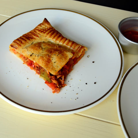 Tuna placed in puff pastry filled with onions, peppers and garlic. On a white plate with a black trim on a cream-coloured table.