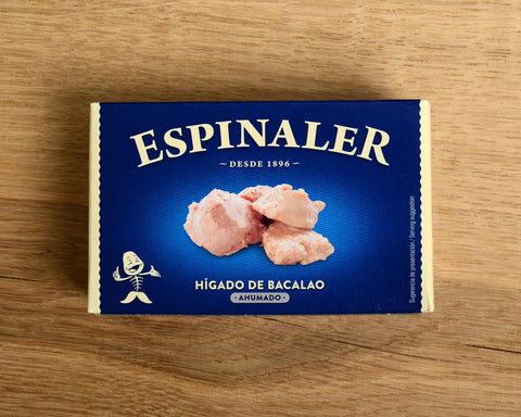 A tin of cod liver in blue card packaging. Espinaler is written in cream lettering above an image of some chunks of cod liver below .