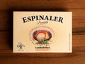 A cream-coloured rectangular box with an open scallop in its shell on the front, beneath Espinaler written in black lettering. The tin is on a wood background.