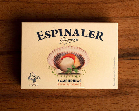 A cream-coloured rectangular box with an open scallop in its shell on the front, beneath Espinaler written in black lettering. The tin is on a wood background.