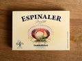 A cream-coloured rectangular box with an open variegated scallop in its shell on the front, beneath Espinaler written in black lettering. The tin is on a wood background.