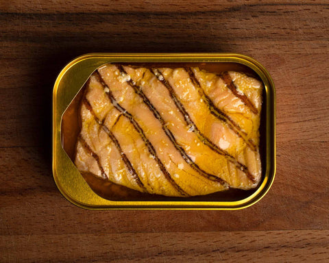 An open tin of salmon with griddle stripes on the fish. The tin is gold against a wood background.