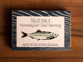 A rectangular box with a blue border and a painting of a herring on the front, against a wood background