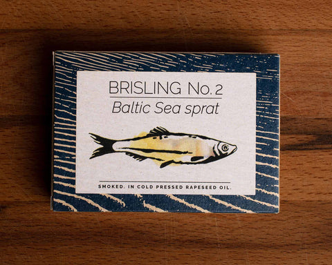 A tin of sprats in a rectangular box with a blue border and a painting of a yellow sprat on the front, against a dark wood background.