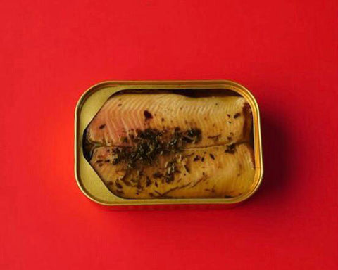 An open golden tin of smoked trout garnished with juniper and lemon thyme, against a bright red backdrop.