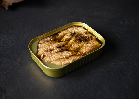 Golden tin of Scandinavian sprats with heather and chamomile. There are around 12 fishes in oil with spices on top. The tin is on top of a black wooden board. 