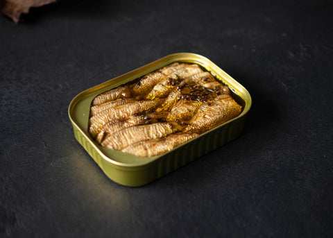 Golden tin of Scandinavian sprats with heather and chamomile. There are around 12 fishes in oil with spices on top. The tin is on top of a black wooden board.