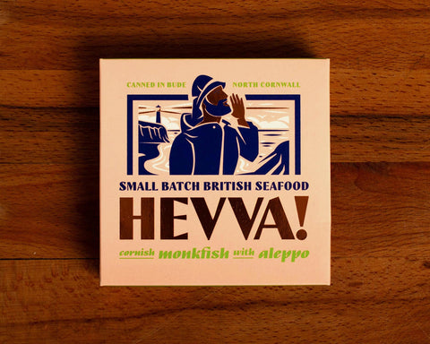 Box containing Cornish monkfish with Aleppo chilli from Hevva! The box is illustrated with a fisherman calling out over the Cornish coast with cliffs and a lighthouse in the background. The box is colourful with Hevva! written in golden, embossed letters