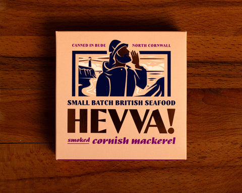 Box containing Cornish mackerel from Hevva! The box is illustrated with a fisherman calling out over the Cornish coast with cliffs and a lighthouse in the background. The box is colourful with the Hevva name written in golden, embossed lettering
