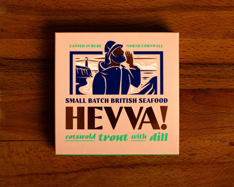 Box containing Cotswold trout with dill from Hevva! The box is illustrated with a fisherman calling out over the Cornish coast with cliffs and a lighthouse in the background. The box is colourful with Hevva! written in golden, embossed letters