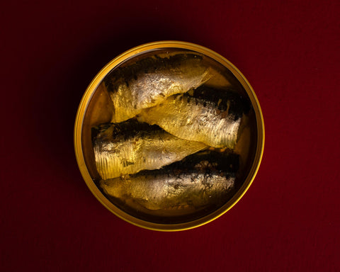 A circular open golden tin of sardines in olive oil, against a dark red background.