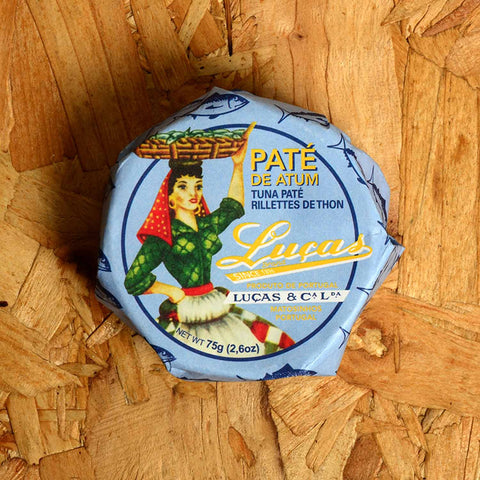 A round tin in a blue paper wrap with Luças written in yellow next to an image of a woman dressed in green and red carrying a basket of fish on her head.