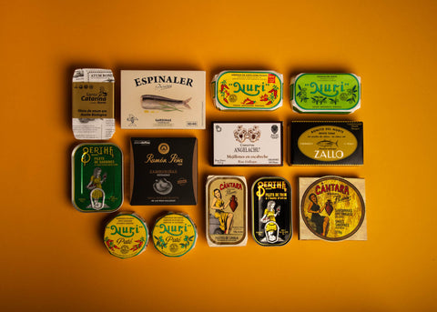 Several tins of Spanish and Portuguese tinned fish, two pâtés and a tin of extra spicy sardines arranged on a yellow background.