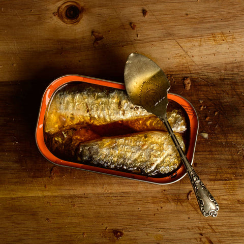 Tinned sardines in a rich orange-tinted oil, on a wooden countertop with a spoon balanced on the tin at the ready.