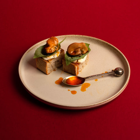 Pepus mussels on crackers with cream cheese and basil leaves placed between them. On a white plate with a vibrant red backdrop.