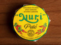 A round tin in a yellow paper wrap. Nuri is written in green lettering above some drawings of red chilli peppers.