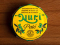 A round tin of sardine pate in a yellow paper wrap. Nuri is written in green lettering above some drawings of olives.