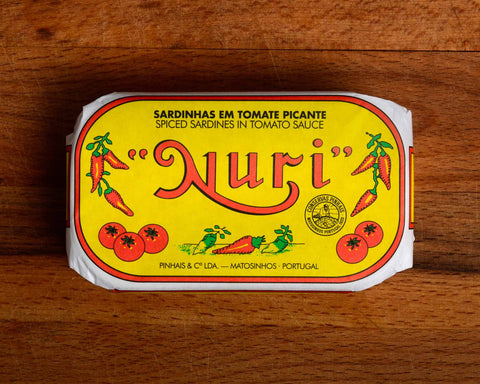 A tin of sardines in spicy tomato in a yellow paper wrap with a white and red border. Nuri is written in red above images of tomatoes and red chilli peppers.