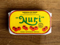 A tin of sardines in a yellow paper wrap with the Nuri logo in green on the front above some images of tomatoes. The tin has a red and white border and is on a wood backdrop.