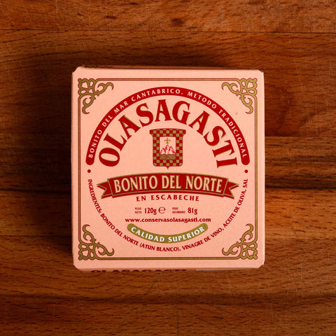 A tin of Bonito del Norte tuna in pink packaging with red lettering and an ornate gold border, against a wood background