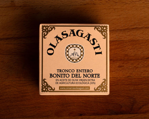 A square tin of tuna with an ornate gold border and Olasagasti written in black lettering against a pale pink background