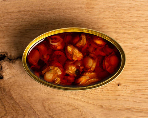 An open, oval-shaped tin of scallops in escabeche against a pale pine wood background.