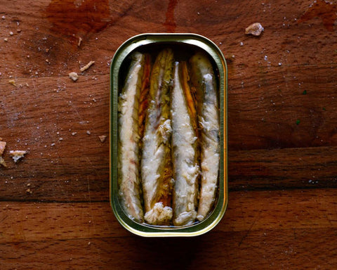An open tin of mackerel fillets in olive oil on a dark wood chopping board with some bread crumbs on it.