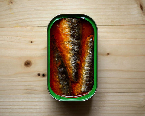 Spicy red tomato flavoured sardines by the tinned fish market. in a green trimmed tin resting on a light brown wooden table
