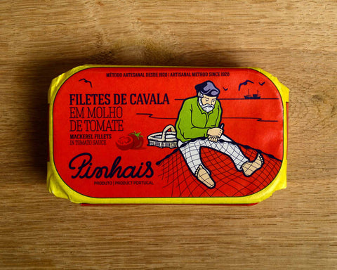 A tin of sardines in a red paper wrap with a yellow border. A fisherman in a green jersey mends his net on the front. The tin is on a wood background.