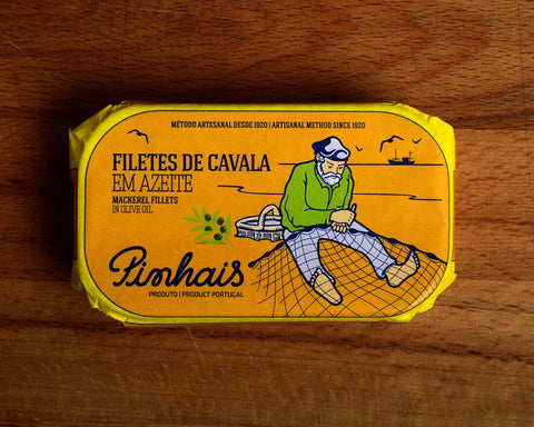 A tin of sardines in orange paper wrap with yellow borders, featuring an illustration of a fisherman with his net on his lap,.Pinhais written in the bottom left corner of the tin.