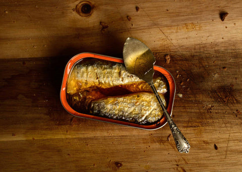 silvery sardines in a bright orange tin with silver spoon on a wooden chopping board with breadcrumbs
