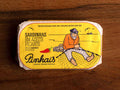 A tin of spicy sardines in a yellow paper wrap with an image of a fisherman mending a fishing net on the front next to a red chilli pepper. The tin is on a wood background.