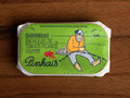 A tin of sardines in a green paper wrap with an image of a fisherman mending a net by some tomatoes and a red chilli pepper. The tin is on a wood backdrop.