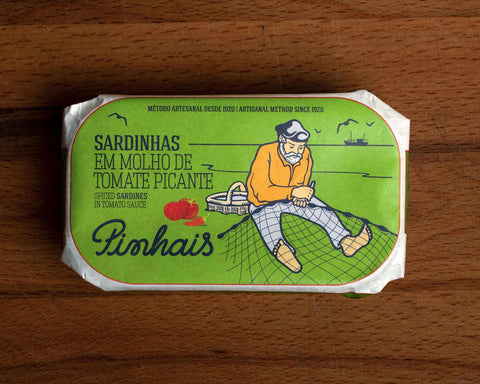 A tin of sardines in a green paper wrap with an image of a fisherman mending a net by some tomatoes and a red chilli pepper. The tin is on a wood backdrop.