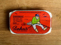 A tin of sardines in a red paper wrap with a white border. A fisherman in a green jersey mends his net on the front. The tin is on a wood background.