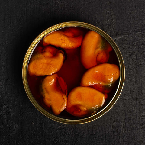 Six large bright orange mussels arranged in a circle inside a circular gold-coloured tin. The tin is against a black background.