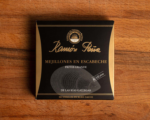A tin of mussels in black card packagaing with a gold border and an image of a mussel drawn on grey on the front. The tin is on a wood background.